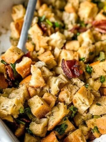 A white baking dish filled with homemade stuffing, featuring cubed bread, herbs, and pecans. A serving spoon rests in the dish. A red cloth is partially visible on the left side.