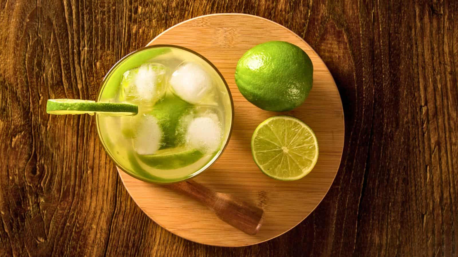 A top-down view of a glass filled with lime wedges and ice cubes on a round wooden board. A whole lime and a half lime are placed next to the glass. The board is on a textured wooden surface.