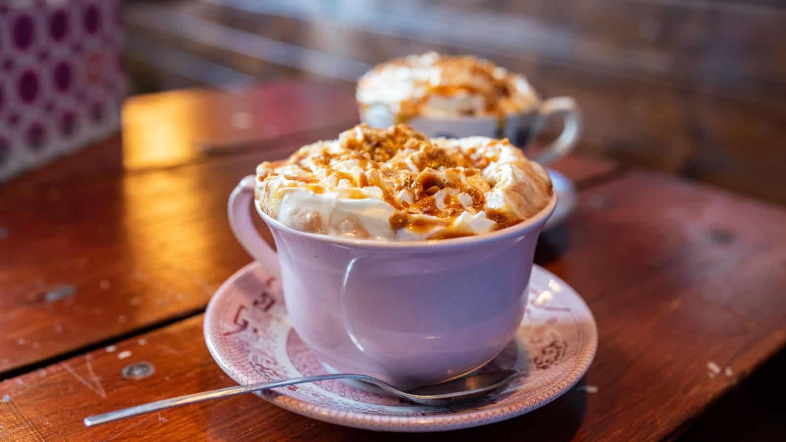A pink mug filled with whipped cream and caramel drizzle sits on a pink saucer with a spoon beside it. The background includes another mug with a similar topping, all placed on a wooden table.
