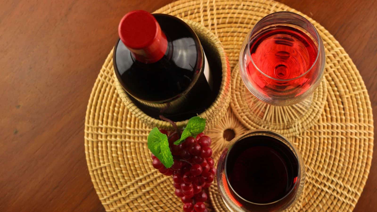 A bottle of red wine and two glasses are placed on a woven mat. One glass contains red wine, while the other is filled with a lighter red liquid, likely ros&eacute;. A small bunch of red grapes is positioned beside the glasses. The surface is wooden.