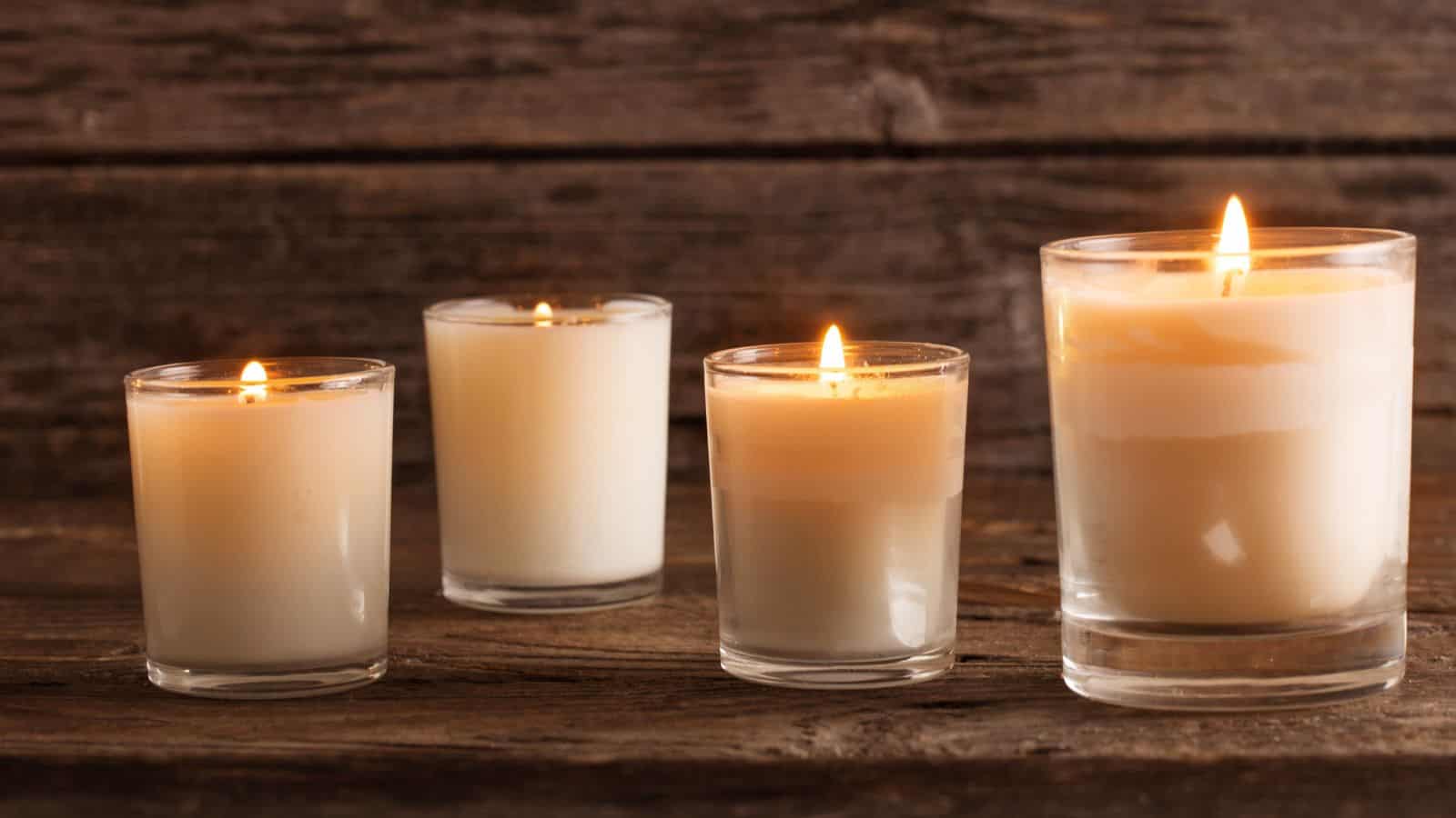 Four lit candles of varying sizes are placed on a wooden surface with a rustic wooden background. Each candle is contained in a clear glass holder, and the flames are gently flickering. The setup creates a warm and cozy atmosphere.