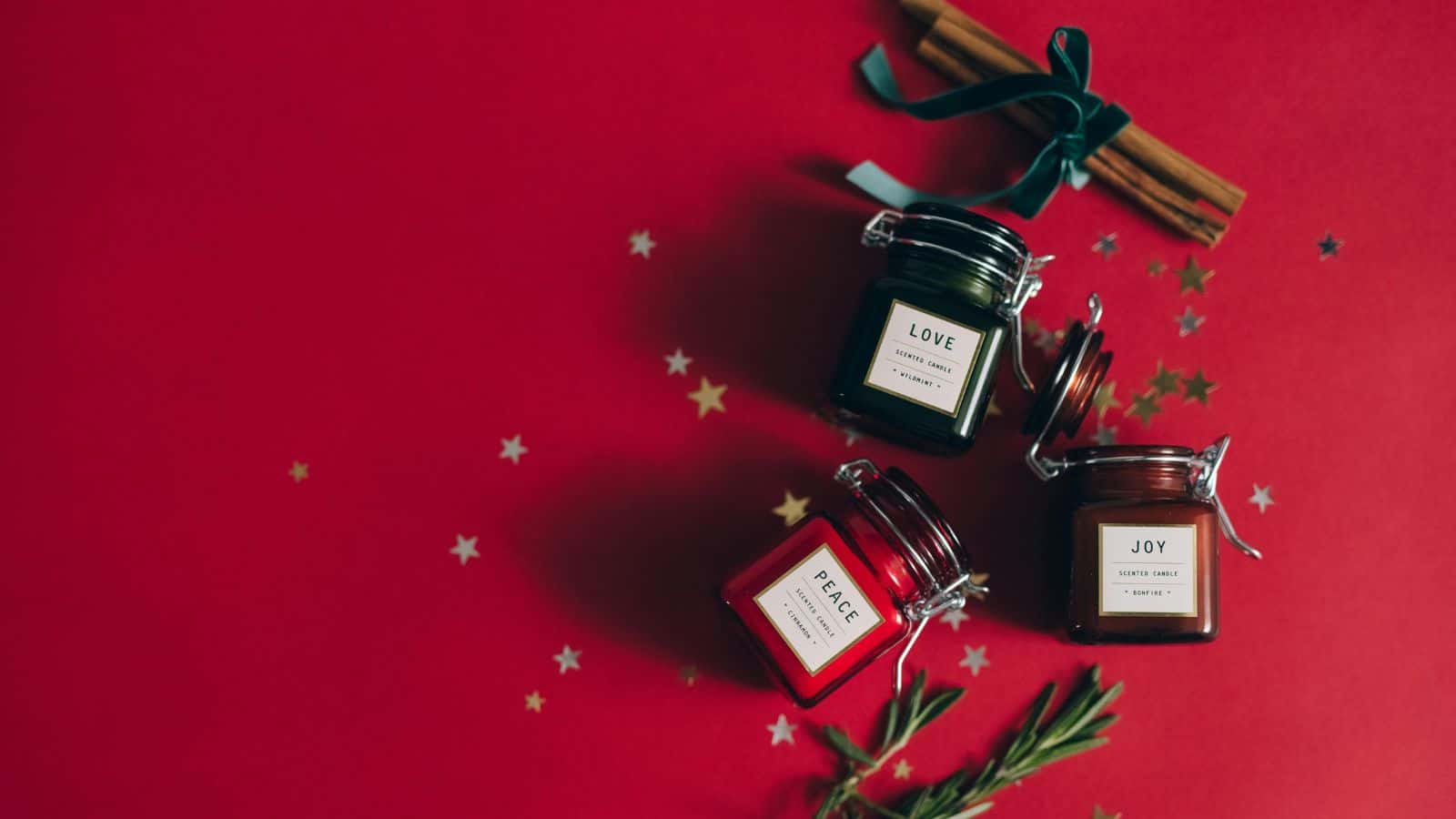 Three small jars labeled "Love," "Peace," and "Joy" are placed on a red surface surrounded by scattered gold stars. Cinnamon sticks tied with a green ribbon and rosemary sprigs are nearby, creating a festive arrangement.