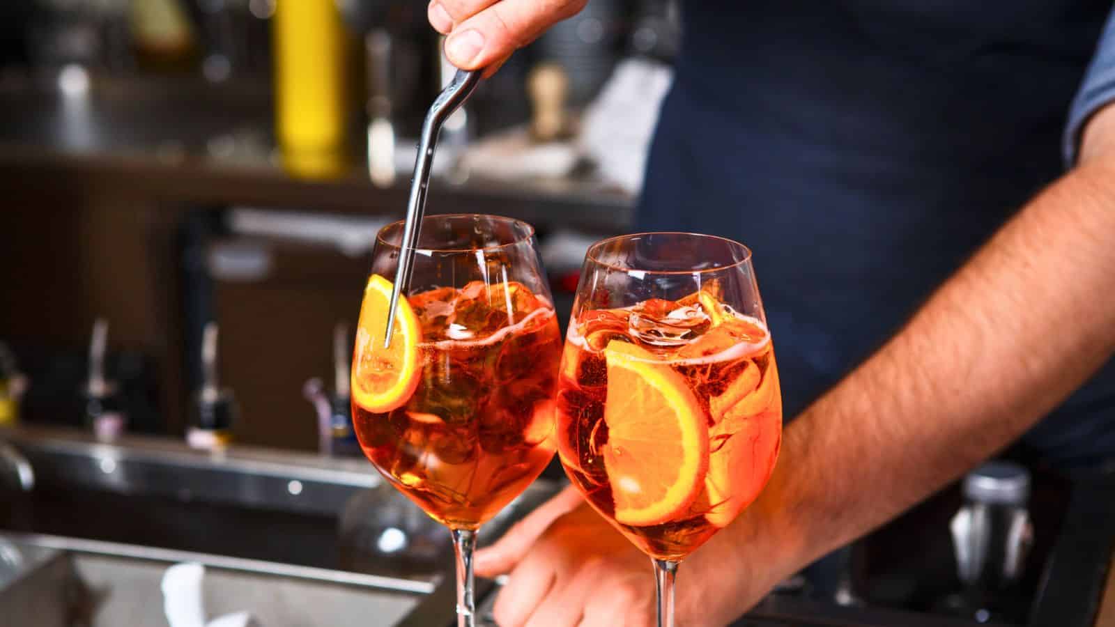 A bartender prepares two glasses of orange cocktail garnished with orange slices, stirring one glass with a metal spoon. The background shows various bar items and bottles.