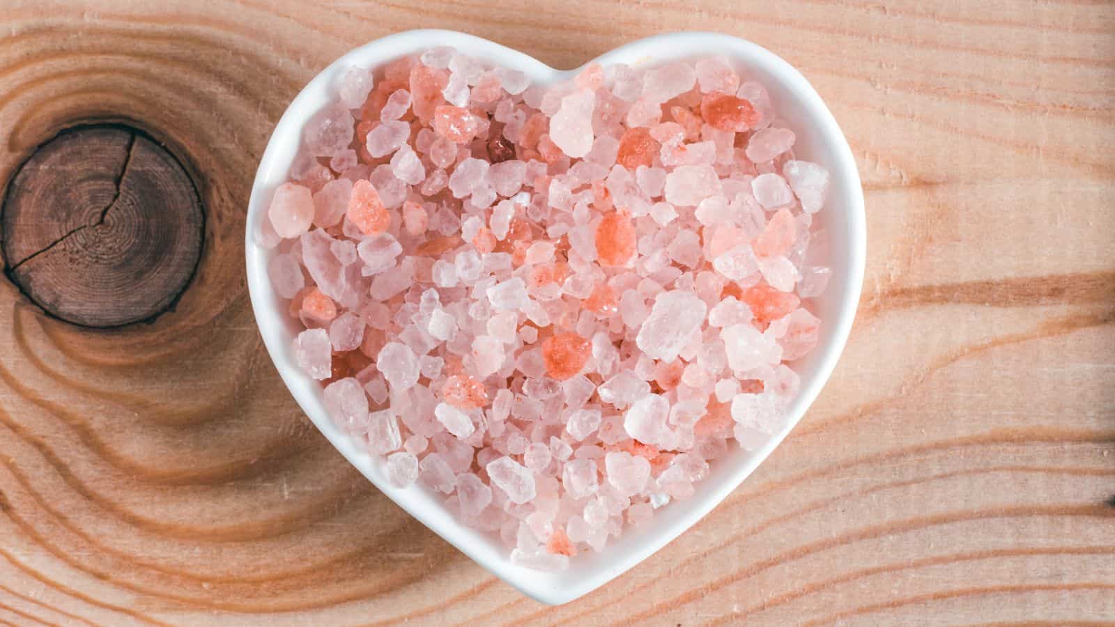 A heart-shaped bowl filled with pink Himalayan salt crystals sits on a wooden surface with visible grain and a knot pattern.