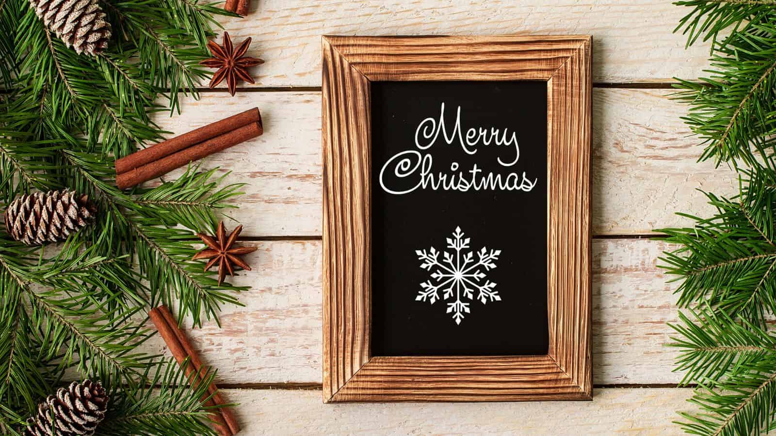 A wooden-framed chalkboard with "Merry Christmas" written in white along with a snowflake sits on a wooden surface. Surrounding it are pine branches, pine cones, cinnamon sticks, and star anise.