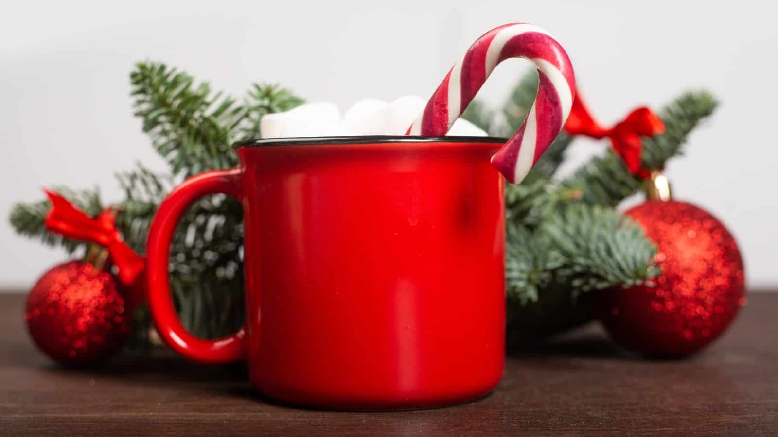 A red mug filled with marshmallows sits on a wooden surface. A candy cane is placed inside the mug. Behind it, there are green pine branches and red Christmas ornaments with ribbons.