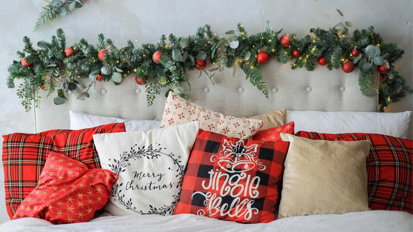 A bed with a headboard adorned with a garland of greenery, red baubles, and lights. The bed has several festive pillows with Christmas designs and patterns, including "Merry Christmas" and "Jingle Bells" text.