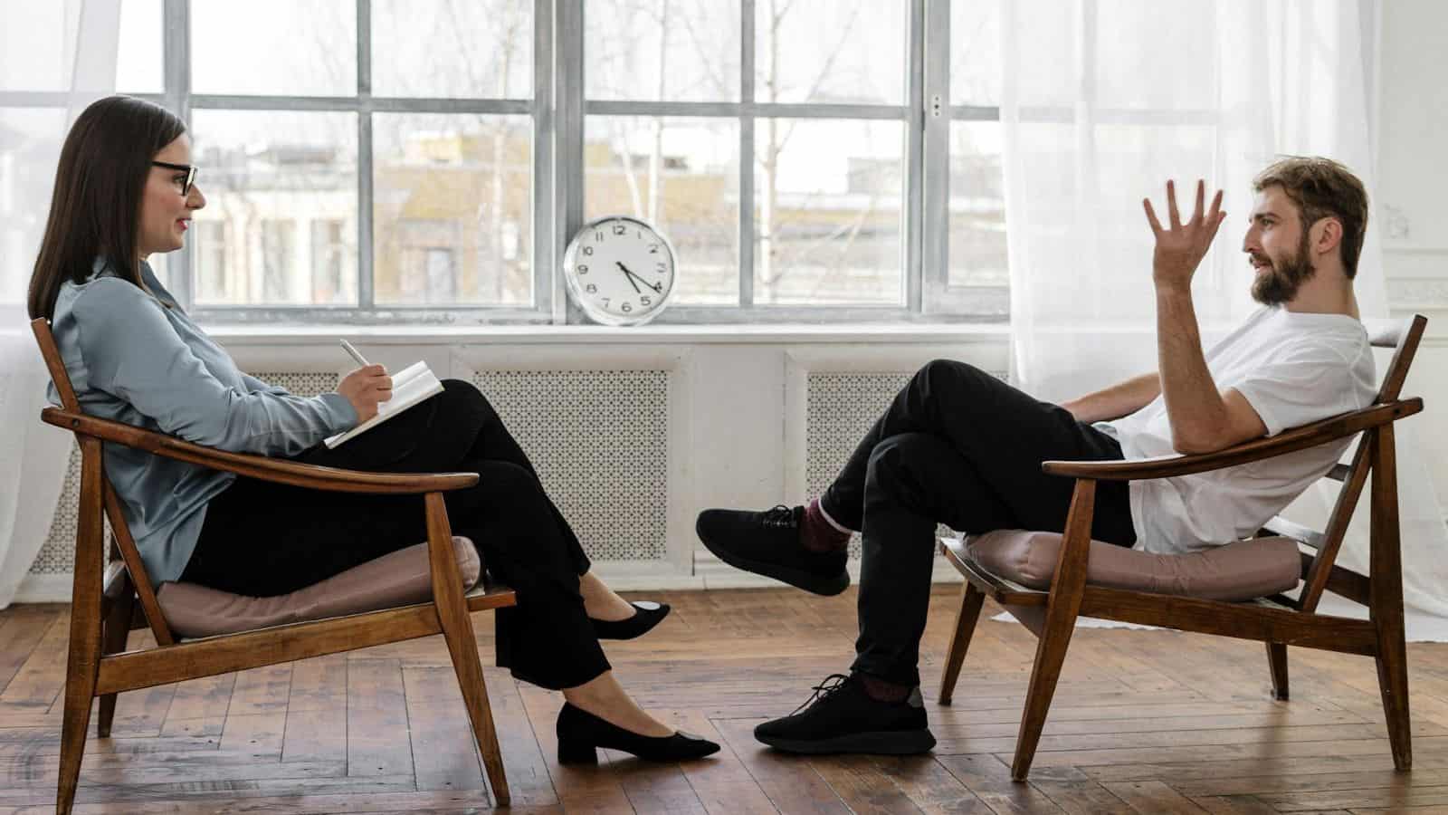 A man and woman are sitting in chairs facing each other in a room with a large window. The man is gesturing with his hand, while the woman is writing on a notepad. A clock is visible in the background, placed on a radiator cover.