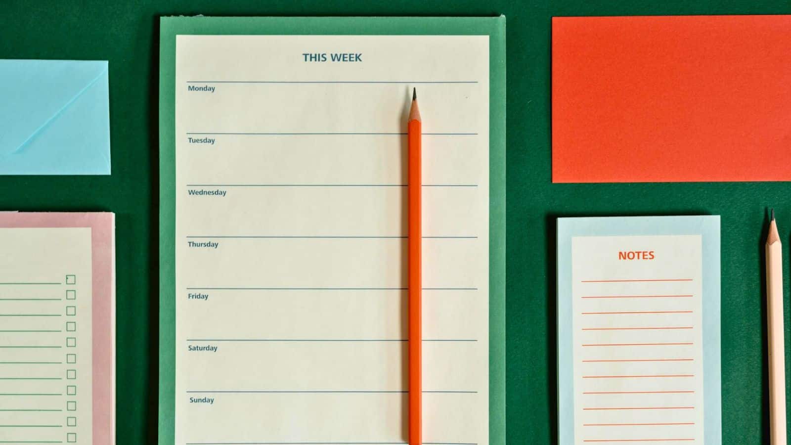 A weekly planner lies on a green surface, featuring days of the week labeled from Monday to Sunday. A red pencil is placed diagonally on the planner. Nearby, there are blank notes, a red envelope, a blue envelope, and a checklist.
