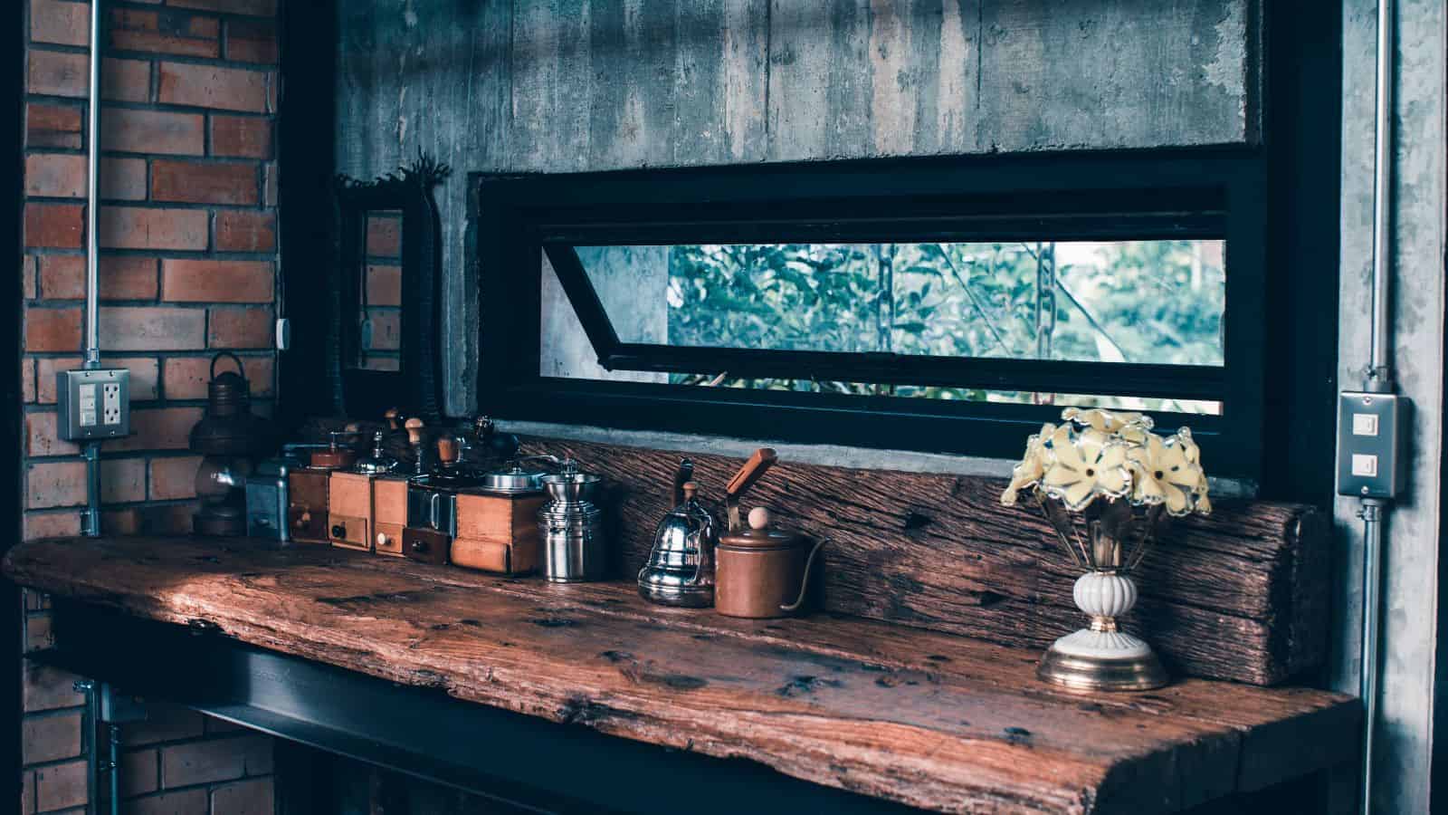 A rustic kitchen counter with wooden beams features a variety of vintage coffee grinders, jars, and a metal kettle. A bouquet of yellow flowers in a decorative vase is placed on the right. A small horizontal window offers a view outside.