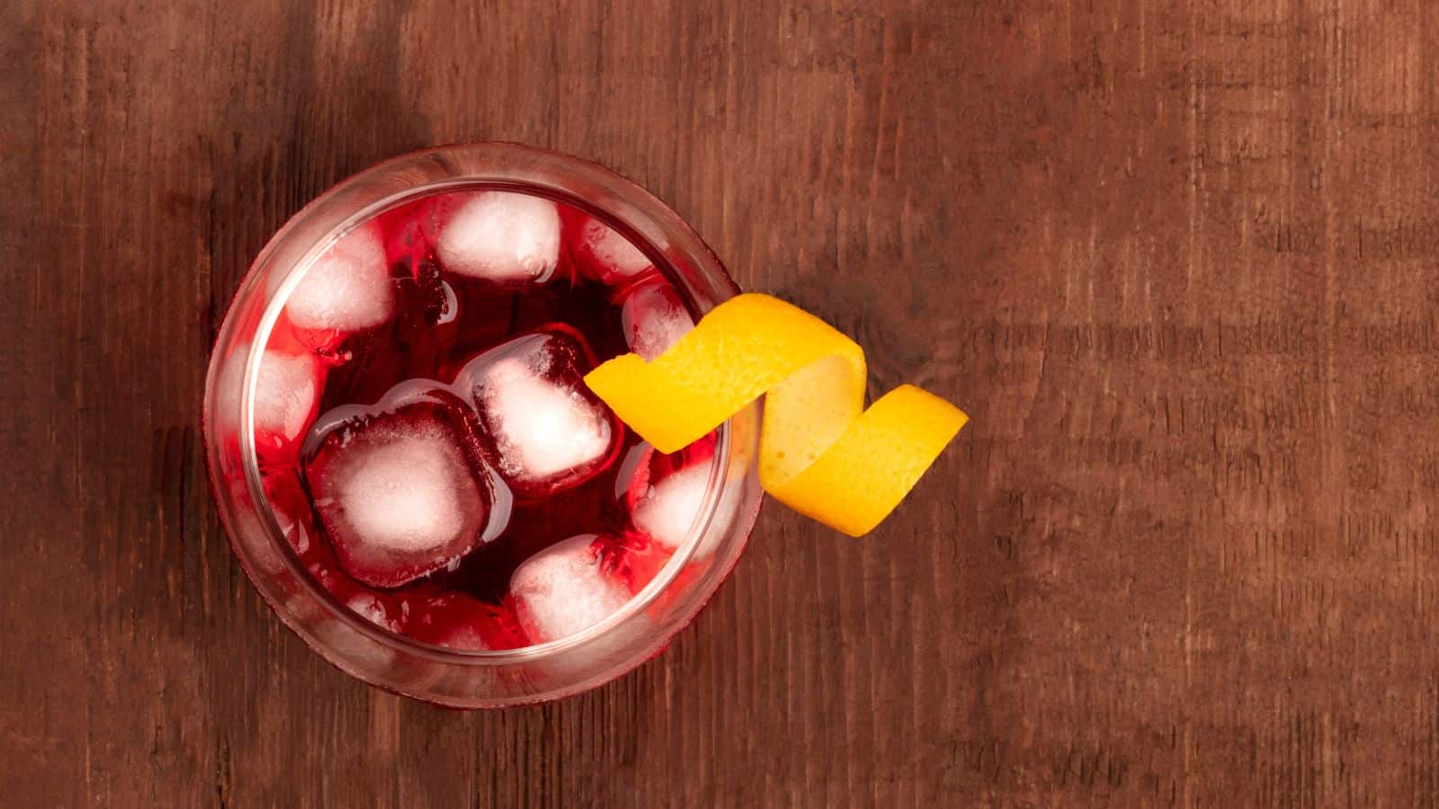 A top-down view of a glass filled with a red cocktail and ice cubes on a wooden surface. A twisted orange peel garnish is placed on the rim of the glass.