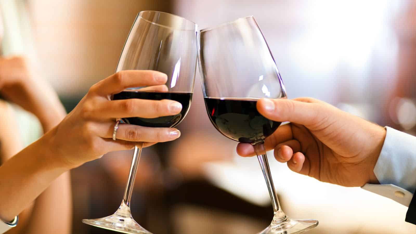 Two hands holding wine glasses filled with red wine are clinking them together. The background is softly blurred, suggesting an indoor setting, possibly a restaurant or caf&eacute;. The focus is on the gesture of the toast.