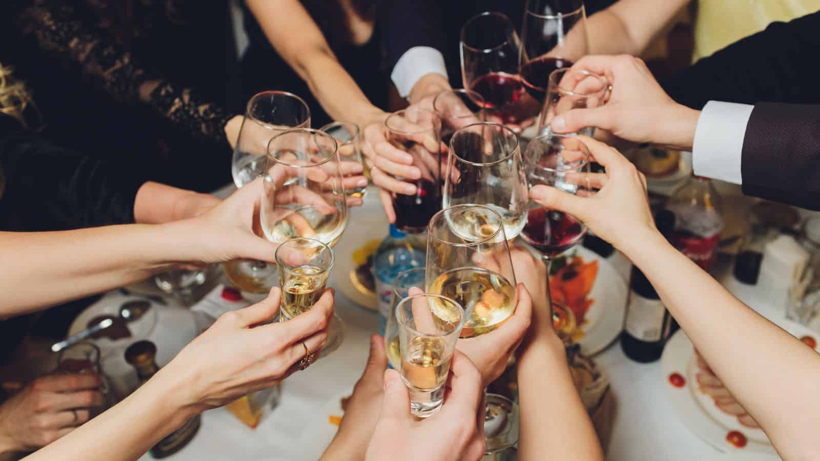 A group of people clinking various glasses, including wine glasses and shot glasses, in a celebratory toast. The table is lightly adorned with bottles and plates of food.