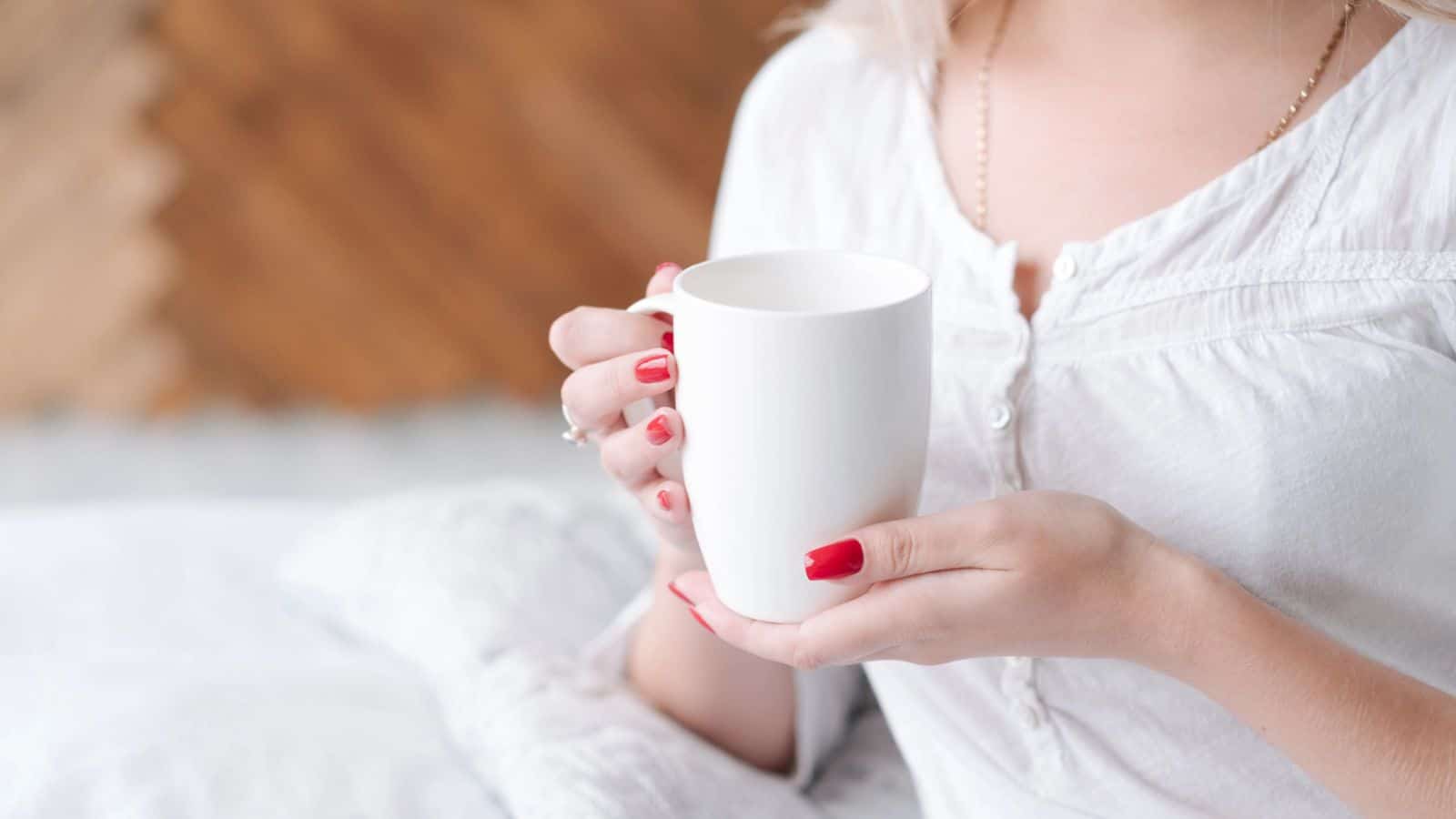 A person with red nail polish holds a white mug with both hands. The person is wearing a white shirt. The background is blurred, showing a hint of a wooden surface.