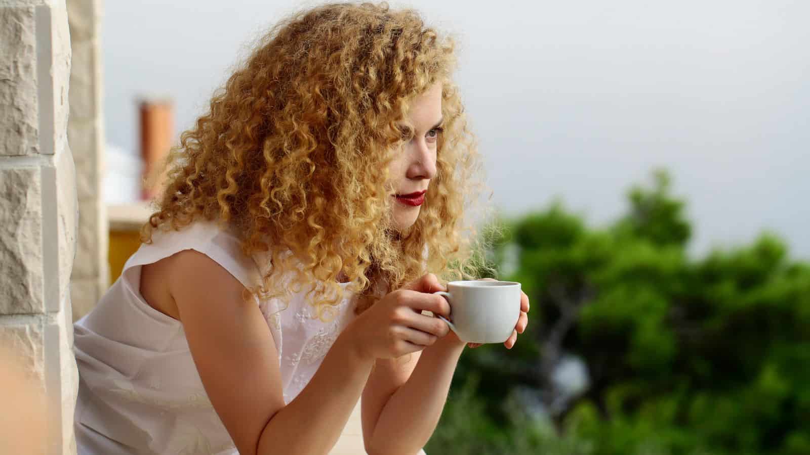A person with curly hair holds a white cup while leaning on a balcony railing. They are wearing a sleeveless white top. In the background, there is greenery and an overcast sky.