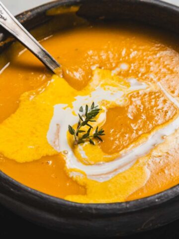 A ceramic bowl filled with creamy orange soup, garnished with a swirl of cream and a sprig of thyme. A spoon rests in the bowl, and it's placed on a dark plate over a marble surface.