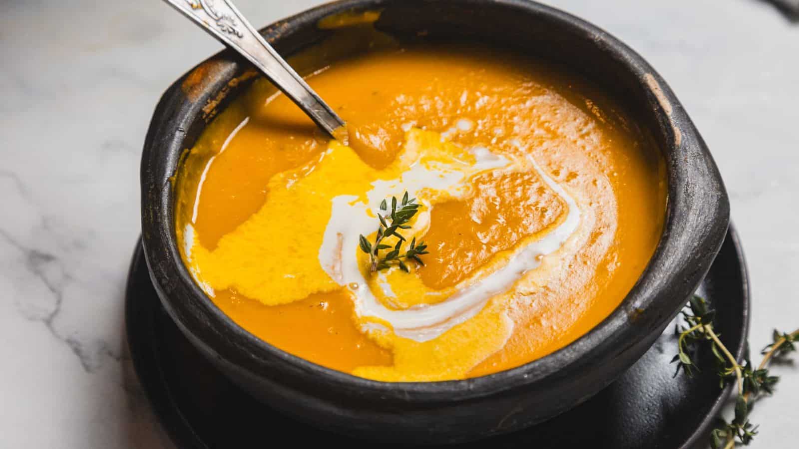 A ceramic bowl filled with creamy orange soup, garnished with a swirl of cream and a sprig of thyme. A spoon rests in the bowl, and it's placed on a dark plate over a marble surface.