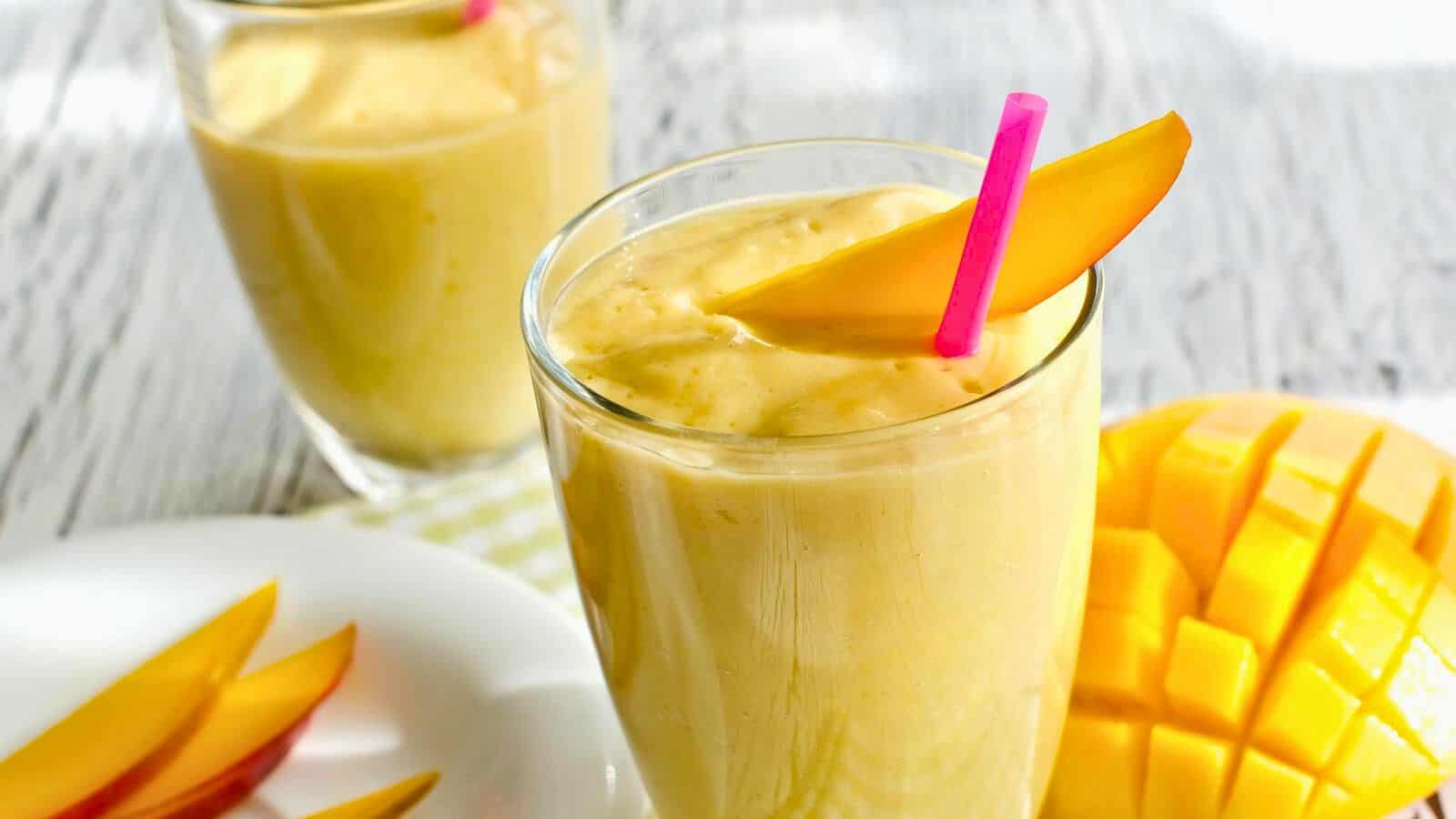 A glass of mango smoothie with a pink straw and a mango slice on the rim is in the foreground. Another glass is in the background. A plate with mango slices and a partially sliced mango are also visible. The setting is a light-colored surface.