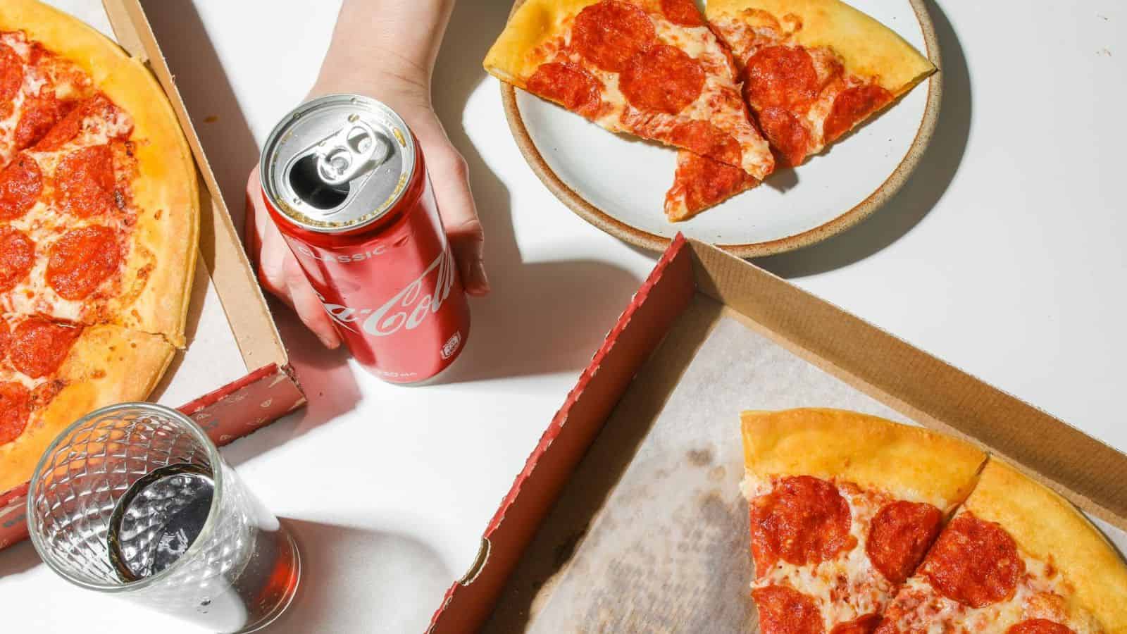 A person holding a can of Coca-Cola sits at a table with two open pizza boxes and a plate containing two slices of pepperoni pizza. A glass of soda is also visible on the table.