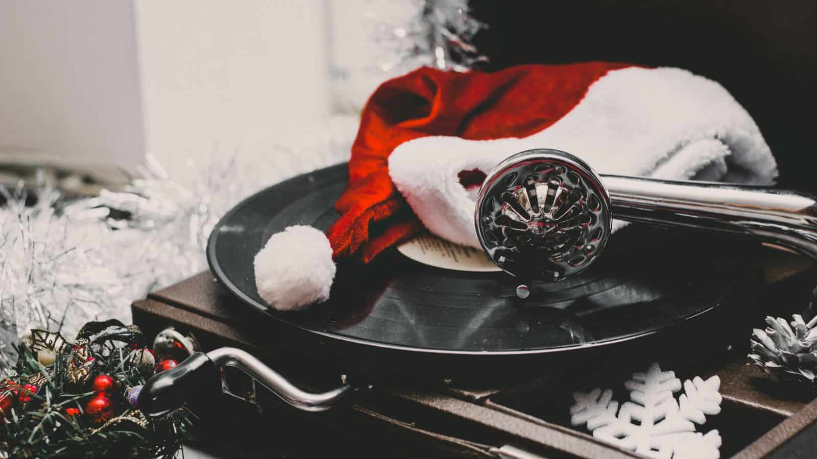 A Santa hat rests on a vinyl record on a turntable, with a decorated wreath and white snowflakes nearby. The scene includes silver tinsel, evoking a festive, holiday atmosphere.