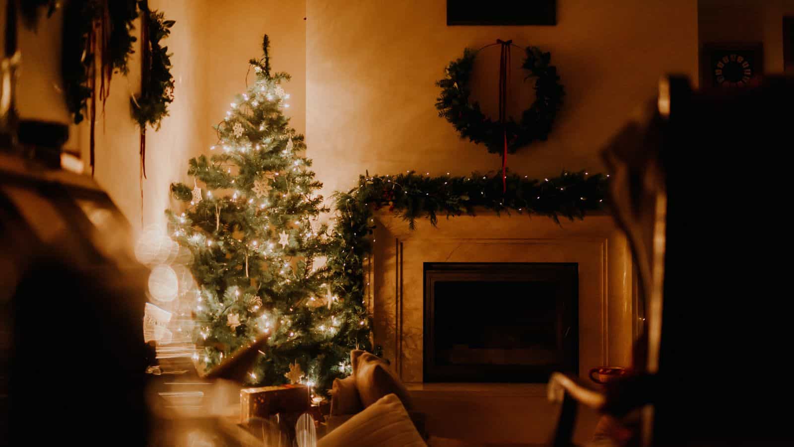 A warmly lit living room features a decorated Christmas tree with lights and ornaments. A wreath hangs above a fireplace, adorned with garlands. The scene conveys a cozy, festive atmosphere.