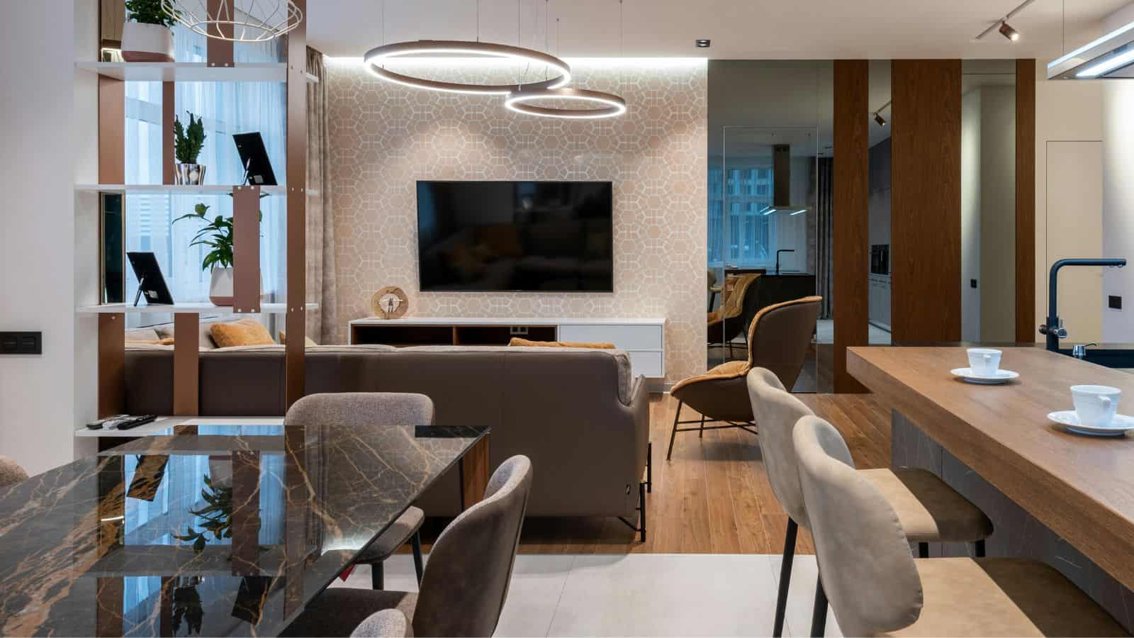 A modern living and dining area with a brown sofa facing a wall-mounted TV. The open space features a marble dining table, bar stools, and a kitchen counter with two cups. Circular light fixtures hang from the ceiling, and plants decorate the shelves.