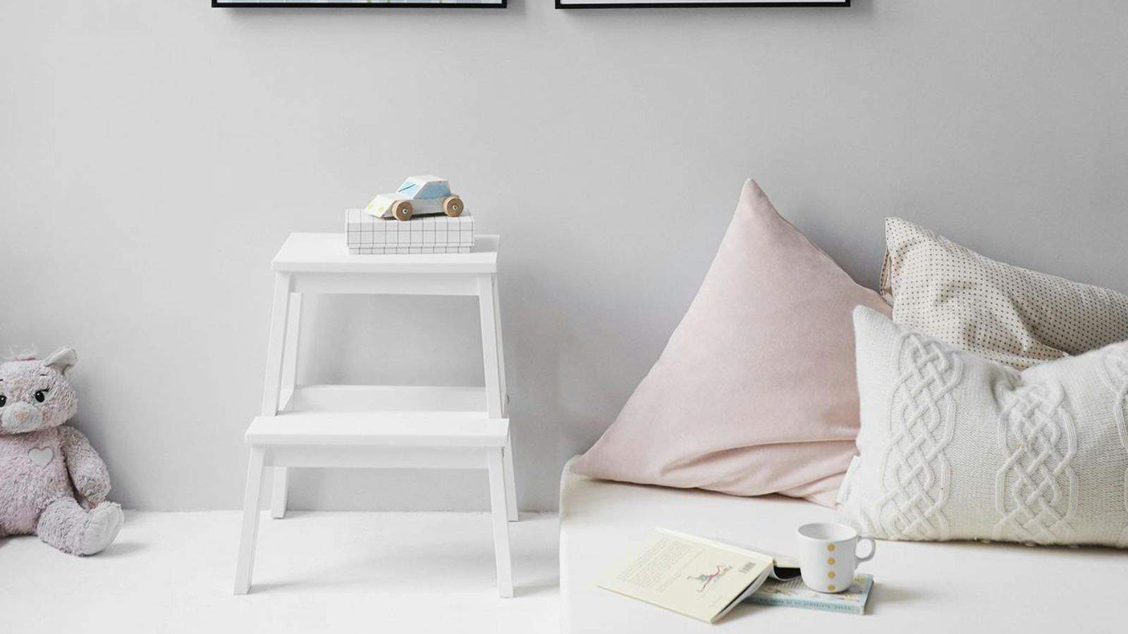 A cozy corner with a small white step stool holding a toy car, soft cushions on a white couch, and a teddy bear on the floor. A polka-dot cup and an open book are placed in front of the cushions. Two framed pictures hang on the wall.