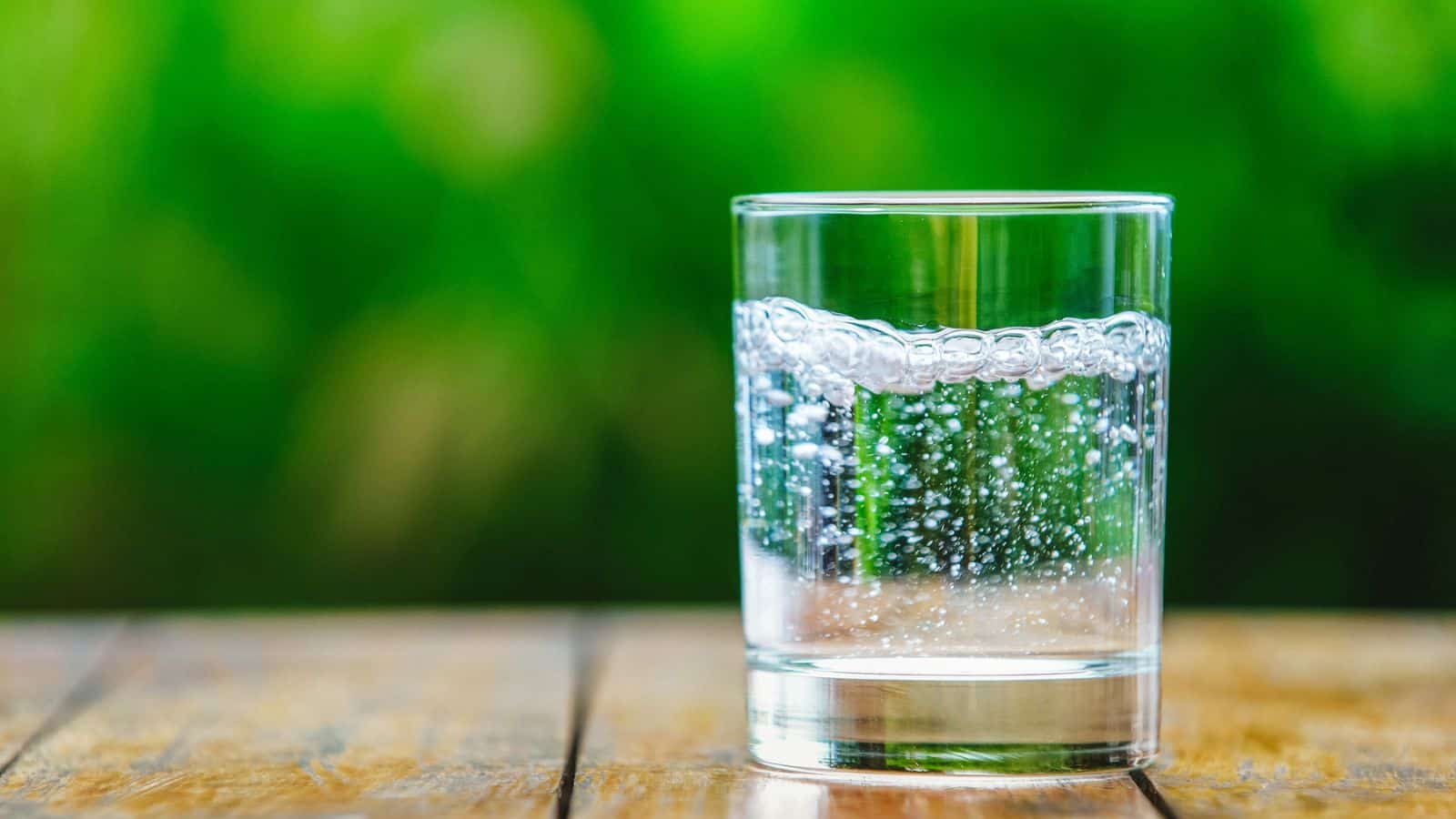 A clear glass filled with sparkling water sits on a wooden surface. The background is a vibrant, blurred green, suggesting a natural or outdoor setting. Bubbles are visible in the water, indicating carbonation.