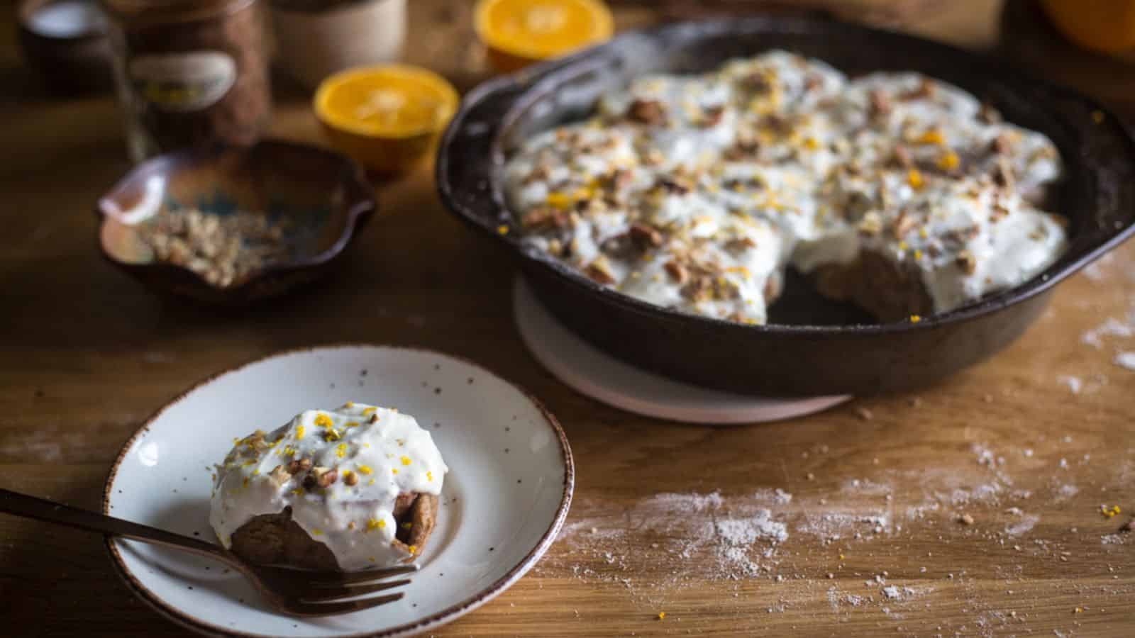 A wooden table with a round pan containing cinnamon rolls topped with cream cheese frosting and orange zest. A single cinnamon roll on a plate with a fork sits beside the pan. Sliced oranges and small jars are visible in the background.