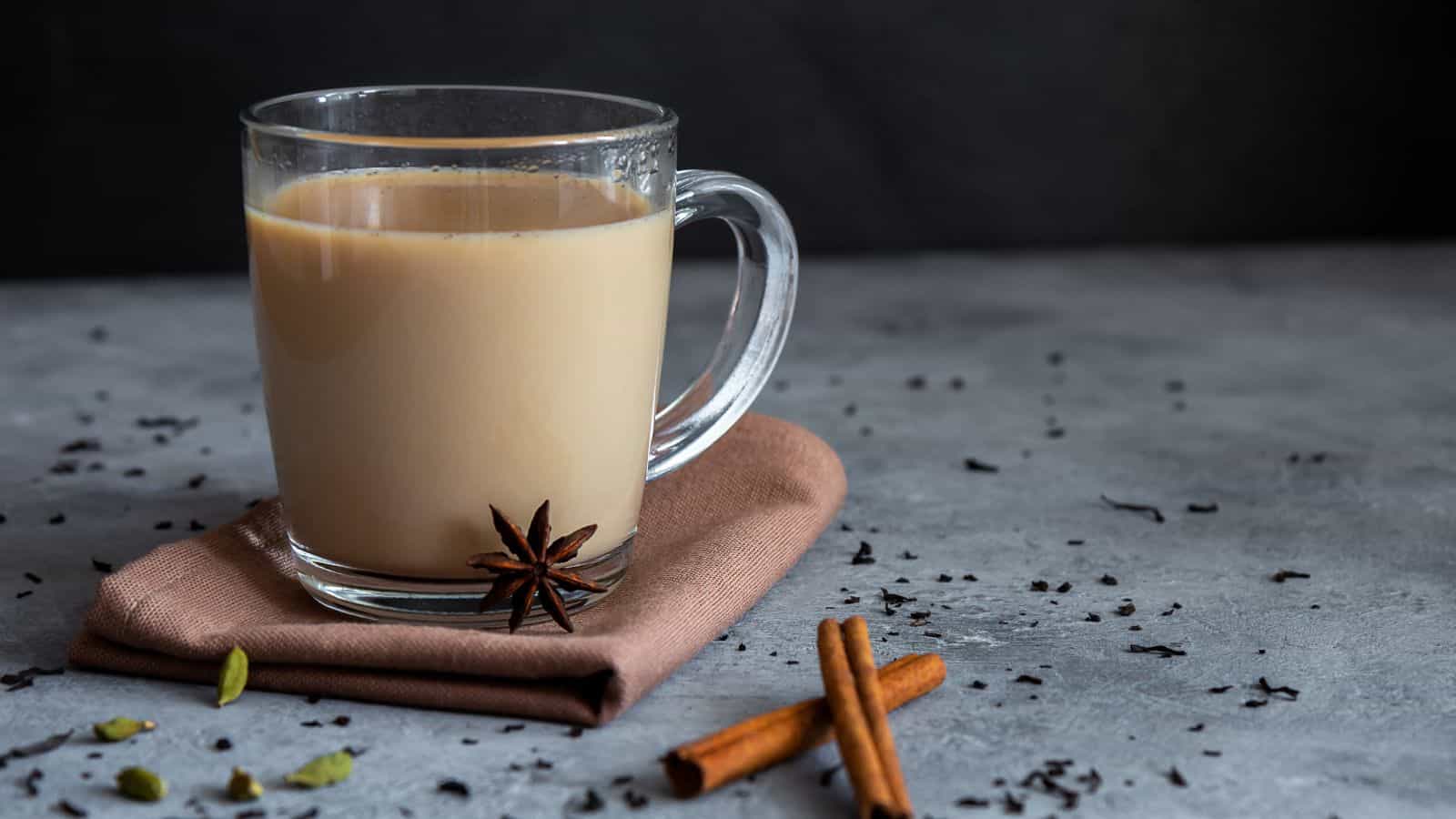 A clear glass mug filled with chai tea sits on a folded beige napkin. Surrounding the mug are scattered tea leaves, cinnamon sticks, star anise, and cardamom pods. The background is a dark, textured surface.