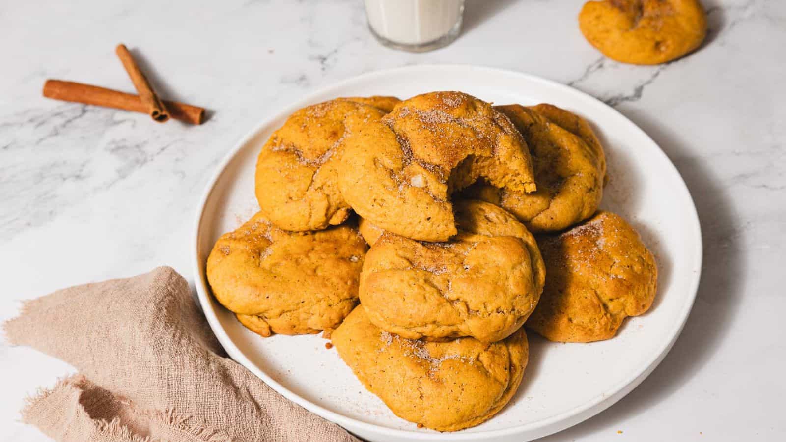 A plate of pumpkin cookies with sugar and cinnamon on top is placed on a marble surface. A bite is taken from one cookie. Two cinnamon sticks and a glass of milk are in the background, along with a beige napkin.