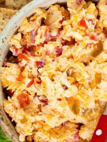 A bowl of pimento cheese spread with a small knife, surrounded by crackers and celery sticks on a white surface. A red and white striped cloth is partially visible on the left.
