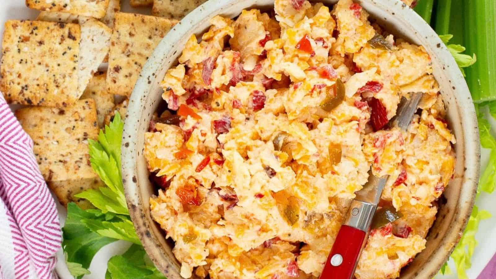 A bowl of pimento cheese spread with a small knife, surrounded by crackers and celery sticks on a white surface. A red and white striped cloth is partially visible on the left.