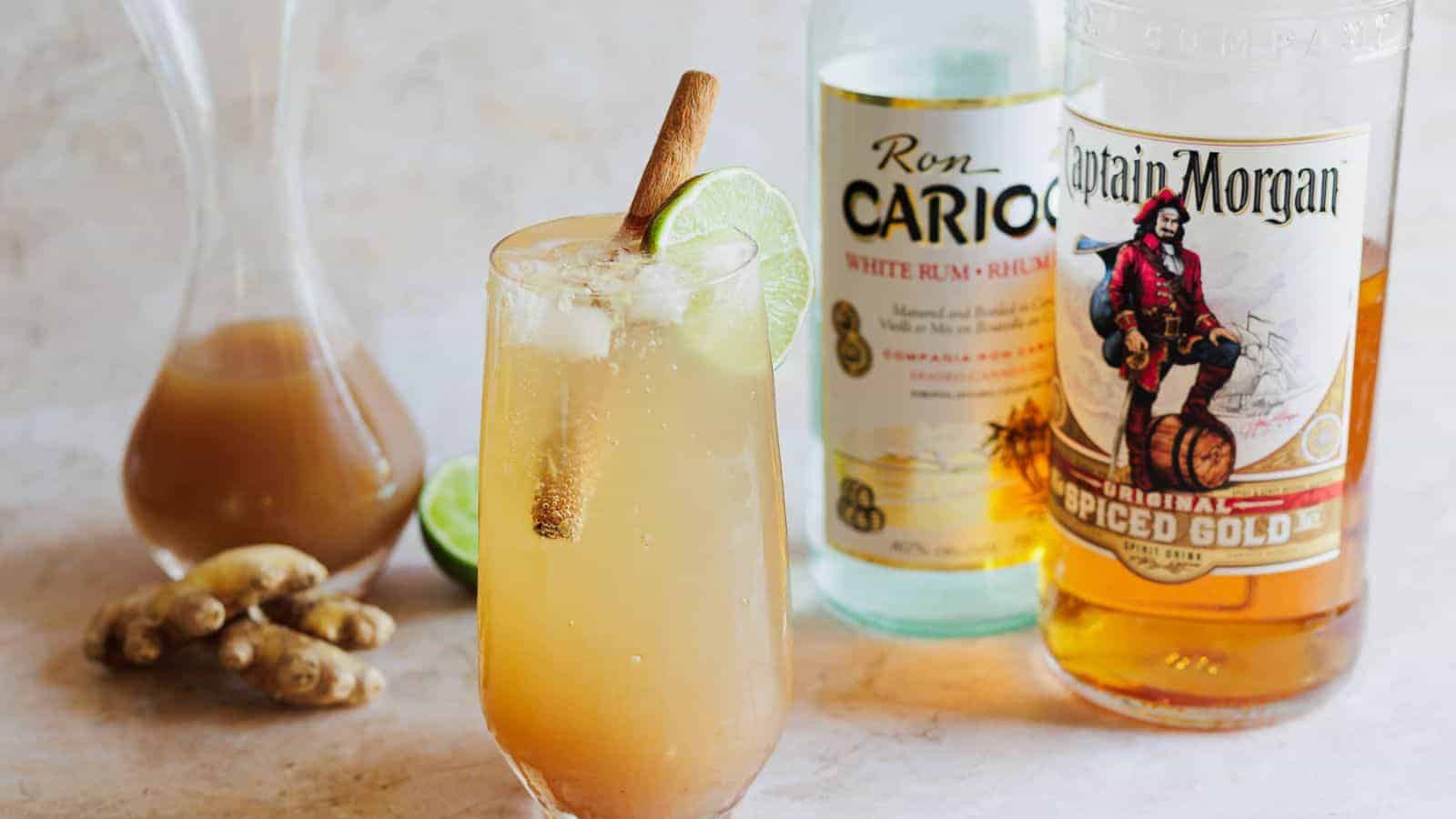A cocktail glass filled with a light brown drink, garnished with a cinnamon stick and lime slice. Behind it are bottles of Ron Carioca white rum and Captain Morgan spiced gold rum. Fresh ginger root and a carafe with liquid are beside them.