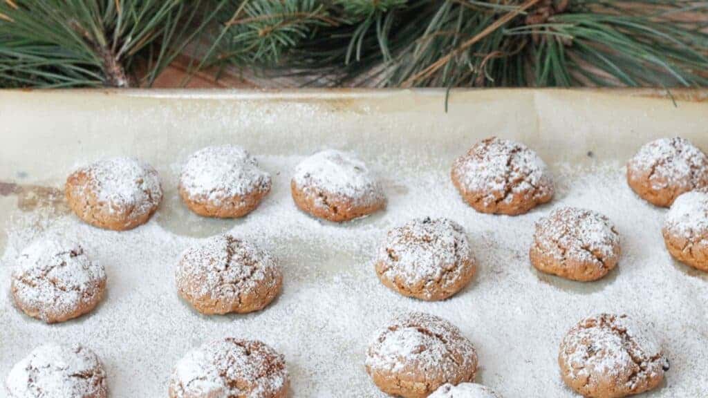 A baking sheet with several round cookies dusted with powdered sugar is displayed. Pine branches are partially visible at the top of the image.