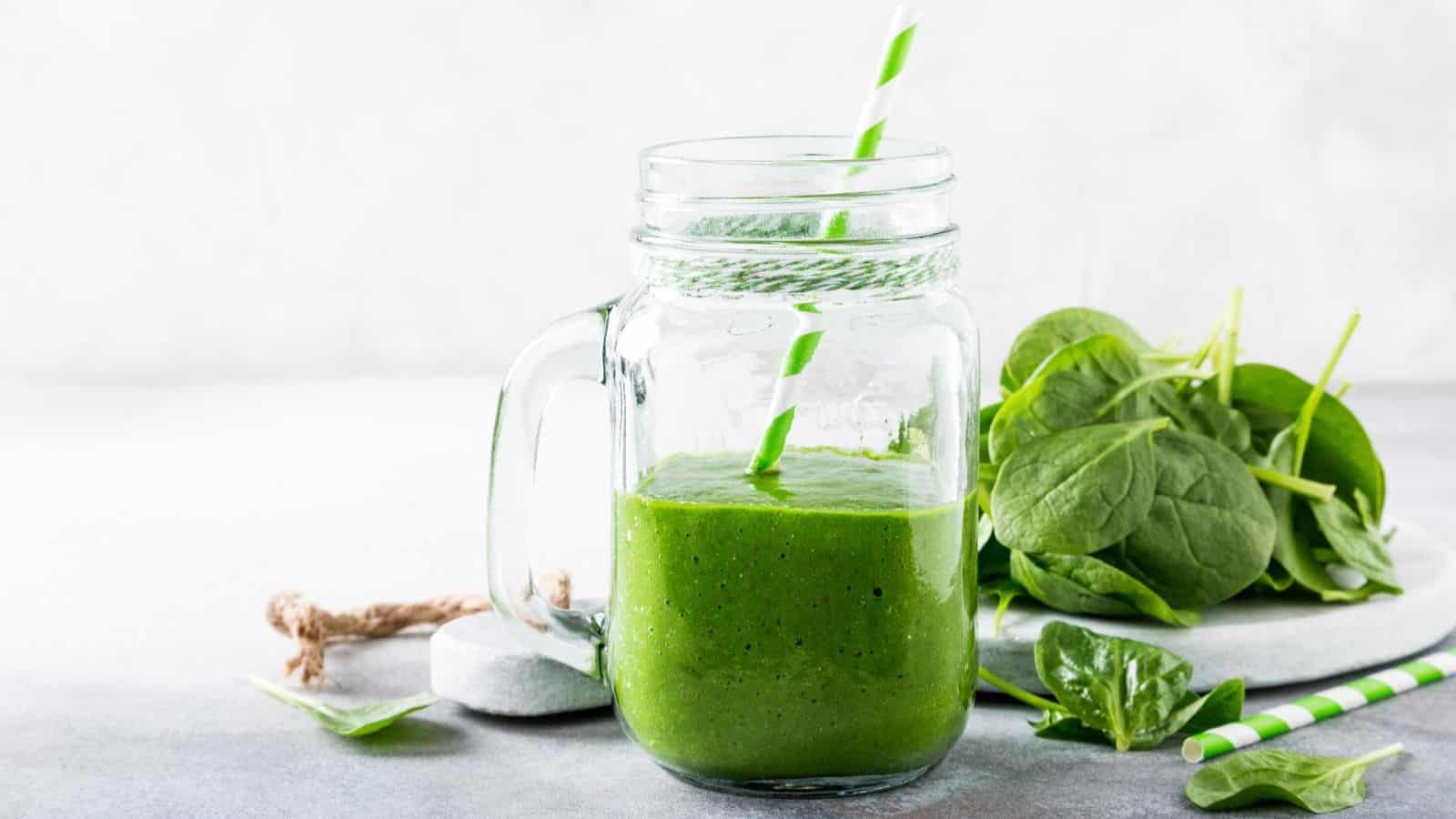 A mason jar filled with green smoothie, featuring a striped straw, is placed on a light surface. Fresh spinach leaves are in the background alongside some scattered leaves. A spool of twine is nearby for decoration.