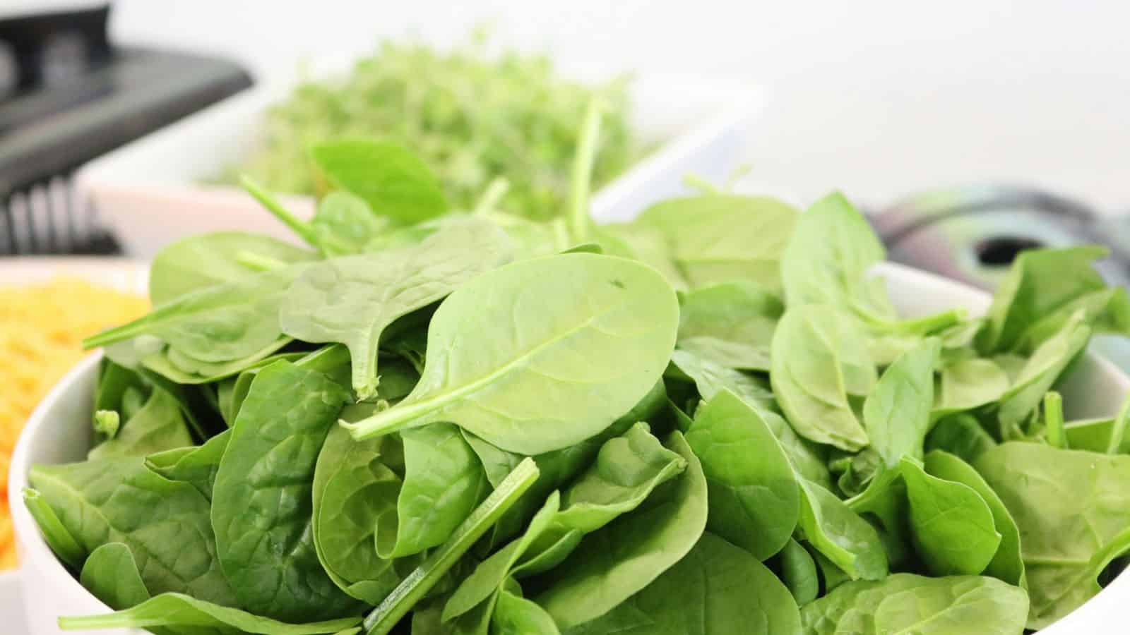 A close-up of a bowl filled with fresh spinach leaves. In the background, there are blurred hints of other ingredients, such as chopped vegetables and greens, on a countertop.