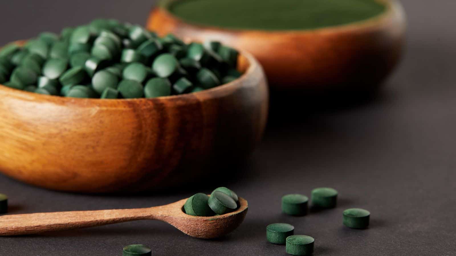 Wooden bowls filled with green tablets and a wooden spoon with tablets on a dark surface.