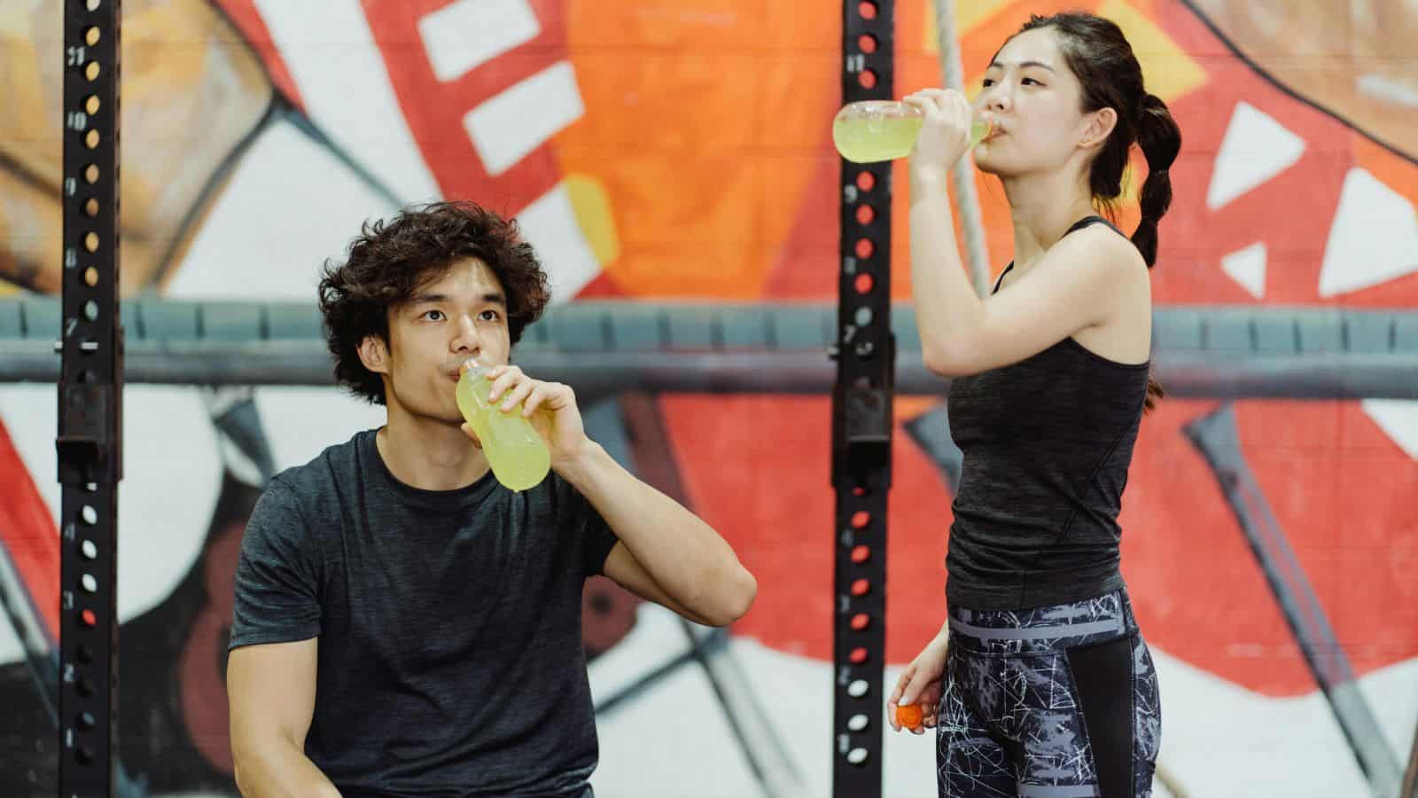 Two people in athletic clothing drink from bottles with a yellow liquid. A seated man and a standing woman pose in front of gym equipment and a colorful mural in the background.