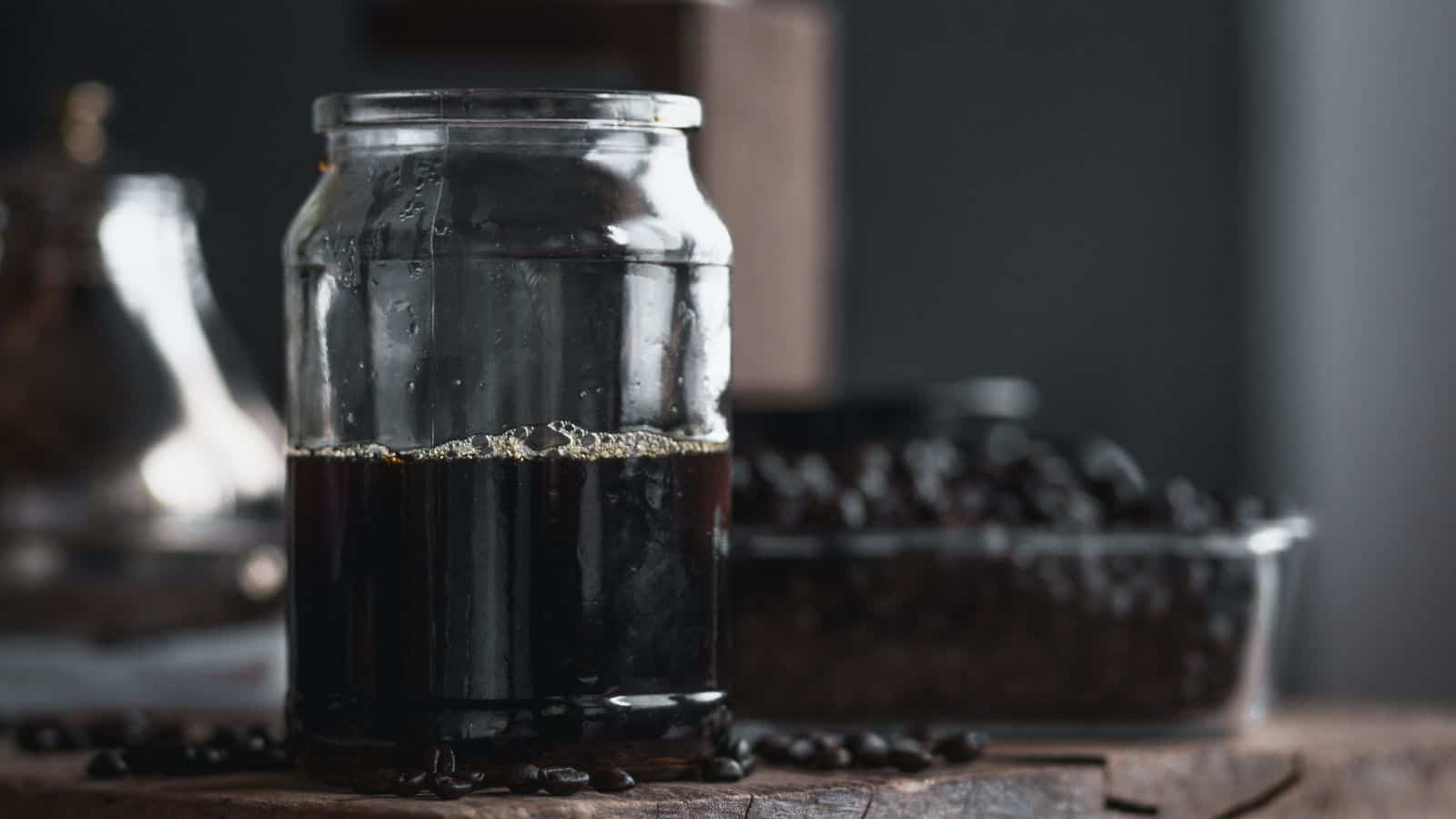 A glass jar filled with dark liquid, possibly coffee, sits on a wooden surface. Coffee beans are scattered nearby, with a blurred container of beans in the background. The lighting is dim and the atmosphere is rustic.