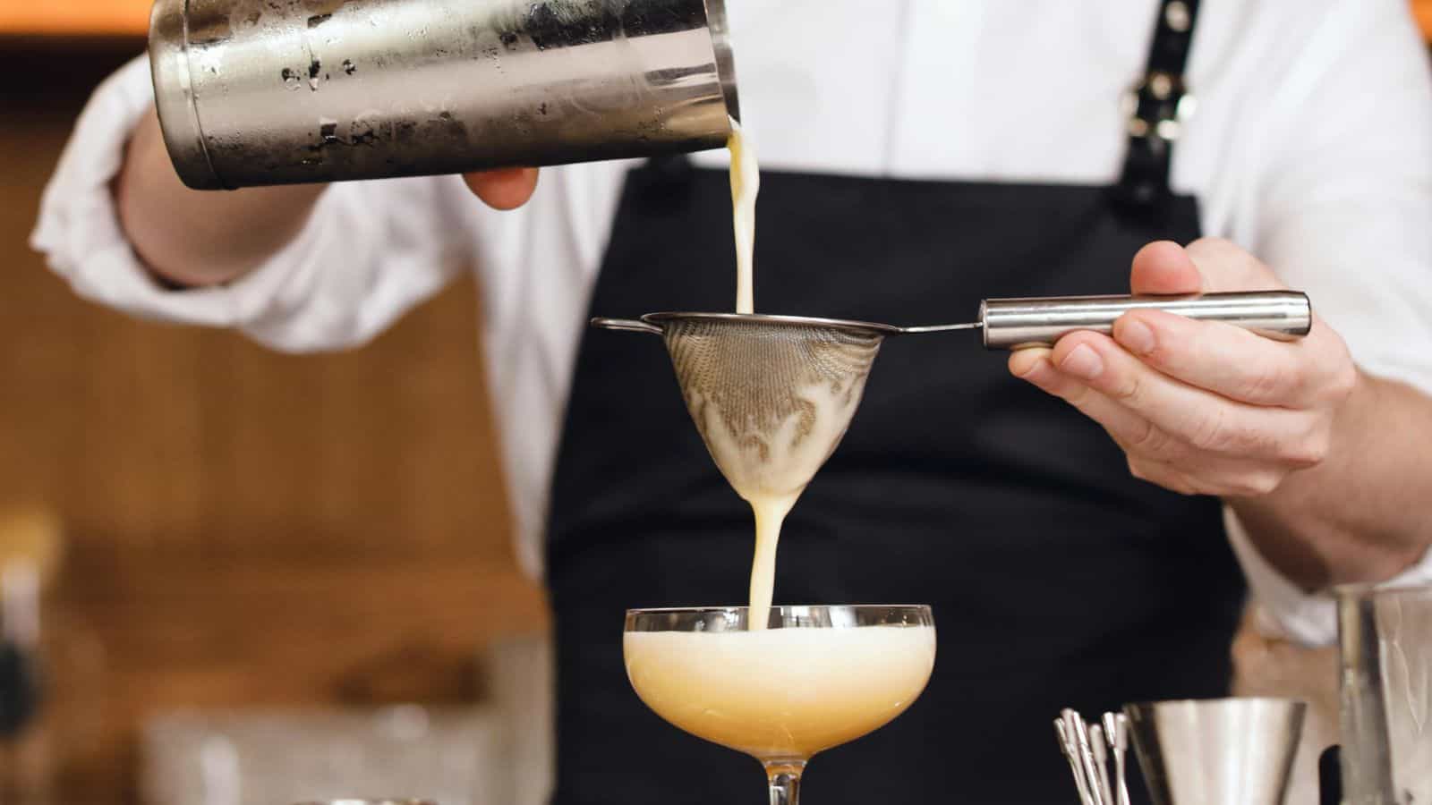 A bartender pours a creamy cocktail through a strainer into a coupe glass. The bartender is wearing a white shirt and black apron. Various bar tools are visible on the counter.