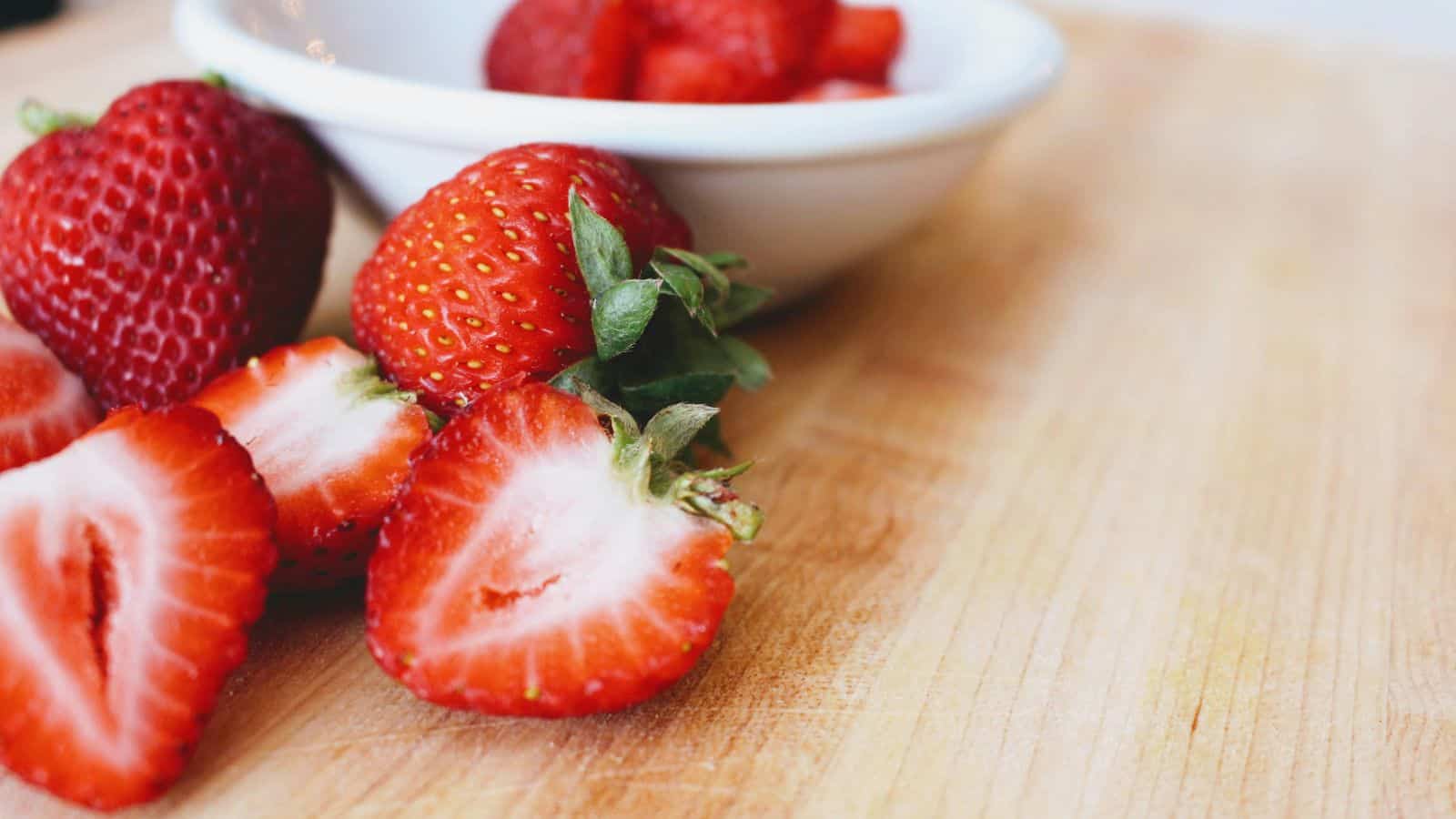 Fresh strawberries, both whole and sliced, are placed on a wooden surface. A white bowl containing more strawberries is in the background. The focus is on the vibrant red color and texture of the fruit.