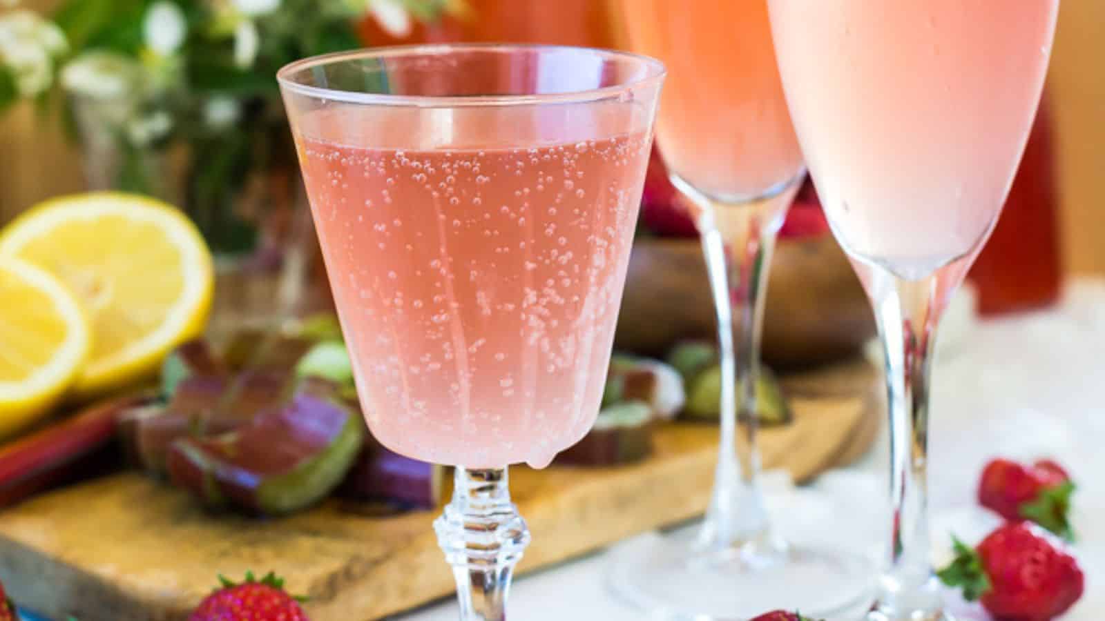 A glass of pink sparkling drink is in focus, surrounded by strawberries, lemon slices, and blurred glasses in the background. A wooden board and flowers are also partially visible in the background.