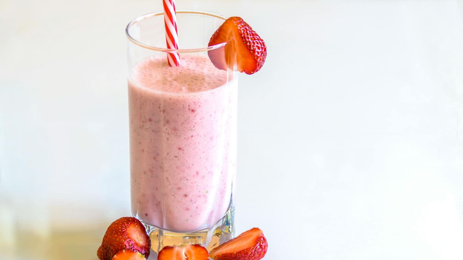 A tall glass of pink strawberry smoothie with a red and white striped straw and a strawberry garnish on the rim. More strawberries are placed around the base of the glass on a white surface.