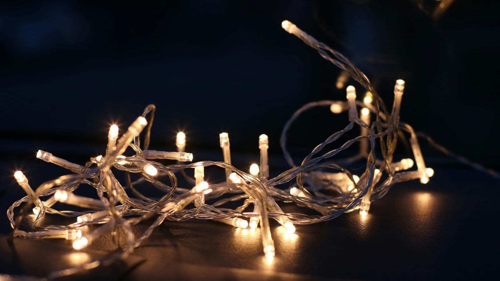 A bundle of warm white LED string lights is illuminated against a dark background. The lights are intertwined, creating a tangled appearance, and emit a soft glow.