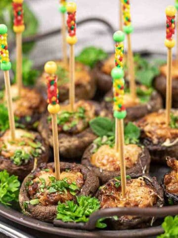 Stuffed mushrooms arranged on a round tray. Each mushroom is topped with cheese and herbs, and is held by a colorful skewer. Fresh parsley is used as garnish around the mushrooms.