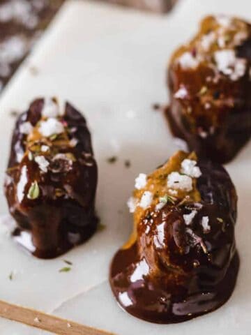 Three dates covered in dark chocolate are placed on a white rectangular marble board. They are sprinkled with sea salt flakes and a few herbs. The board rests on a wooden table.