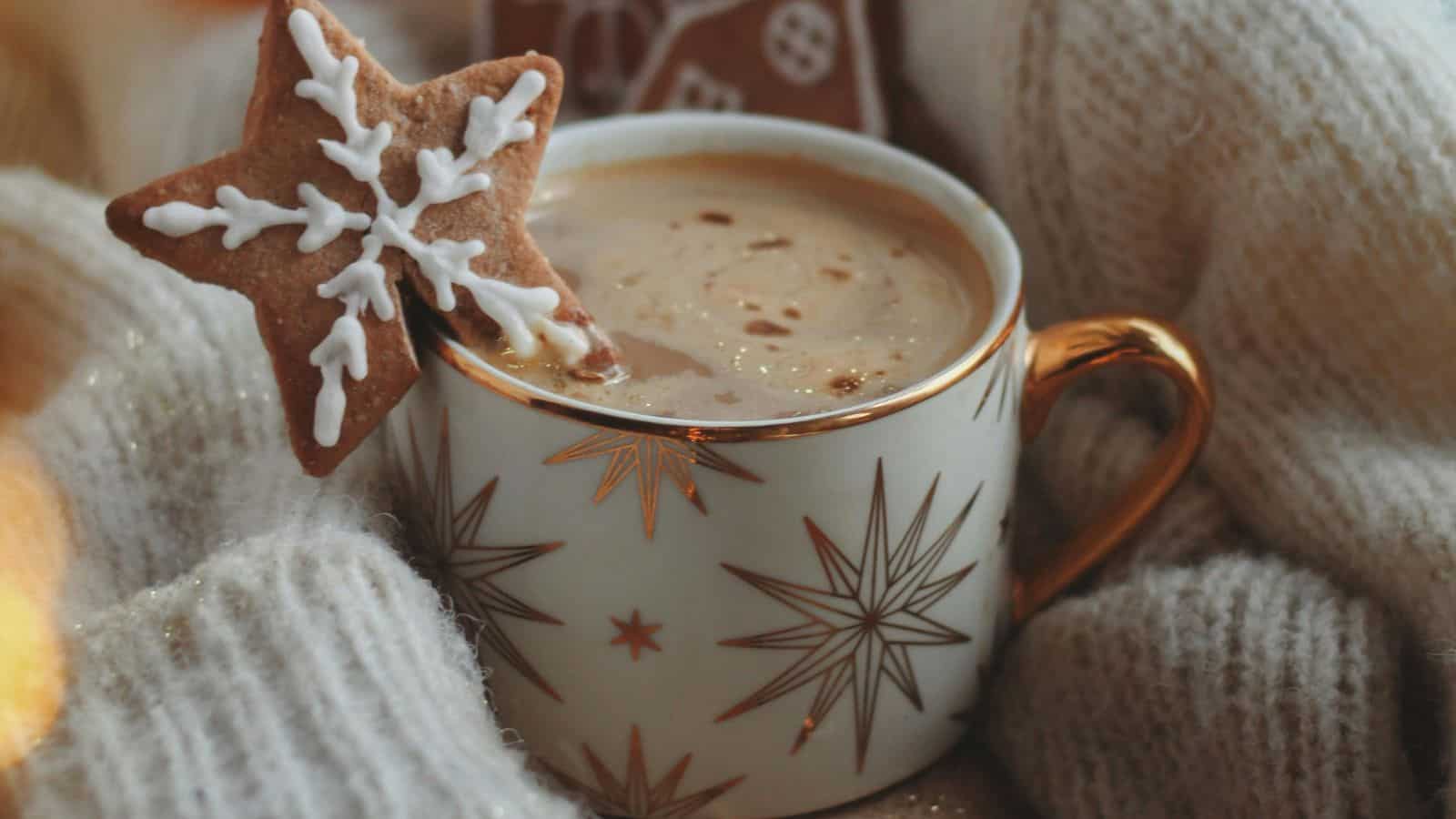 A mug with star patterns is filled with a frothy beverage and topped with a gingerbread star cookie with white icing. The mug is surrounded by soft, cream-colored knit fabric.