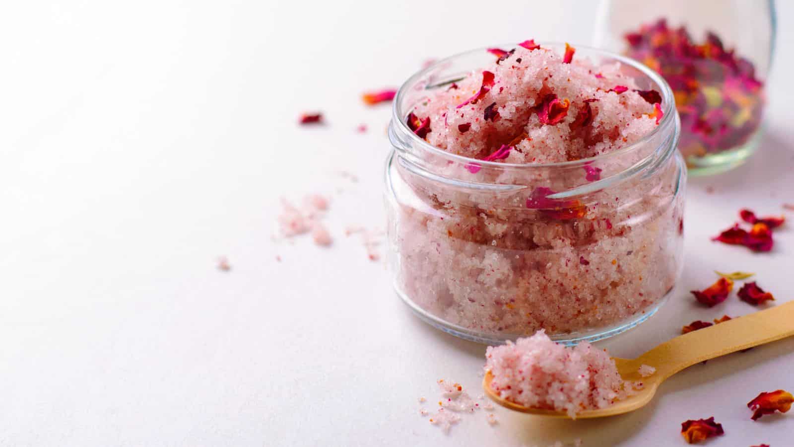 A clear glass jar filled with pink scrub mixed with red petals sits on a white surface. A wooden spoon holds a small amount of the scrub nearby. In the background, another small glass container with more red petals is partially visible.