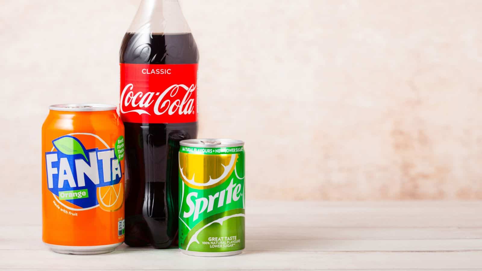 A can of Fanta, a bottle of classic Coca-Cola, and a can of Sprite are placed on a light wooden surface against a beige background.